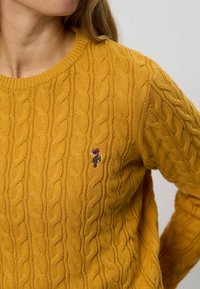 Žena oblečená v horčicovožltej svetri s pleteným vzorom, na hrudi má malú vystuženú nášivku s logom jazdca jazdiaceho na koňovi.