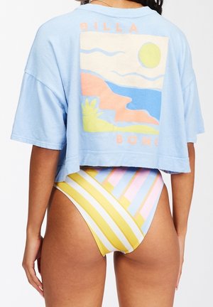 Frau trägt ein hellblaues, kurz geschnittenes T-Shirt mit farbenfrohem Landschaftsprint und hoch taillierte, gestreifte Bikinihosen in Gelb, Pink und Blau.