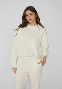 Cremefarbener Hoodie und passende Hose aus weichem Stoff. Verfügt über eine Kapuze mit Kordelzug, überschnittene Schultern und eine lockere Passform mit einer Seitentasche.