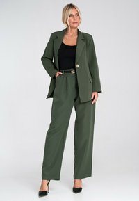 Olivegreenowa marynarka o dopasowanym kroju i szerokie spodnie wykonane z gładkiego materiału, zestawione z czarną bluzką i paskiem z złotymi akcentami. Czarne szpilki.