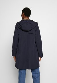 Mörk marinblå vattentät jacka med huva, utrustad med ventilerad rygg, långa ärmar och avslappnad passform. Slät textur, längd till mitten av låret.