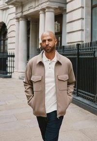 Beige zip-up jakke med to brystlommer, båret over en hvid polo-shirt, parret med mørke jeans. Glat stoftekstur, skræddersyet pasform.