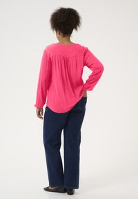 Haut fuchsia à manches longues avec un ourlet incurvé et un tissu texturé, associé à un jean droit bleu foncé et des chaussures plates noires.