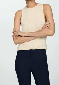 Persona che indossa un top smanicato color crema con pantaloni blu scuro, con le braccia incrociate sul torso davanti a uno sfondo bianco semplice.