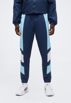 Hombre vistiendo pantalones deportivos azul marino, azul claro y blanco con puños elásticos, combinados con zapatillas blancas Adidas, de pie frente a un fondo liso.