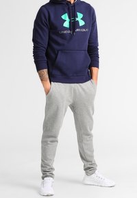 Felpa navy con un grande logo turchese di Under Armour e coulisse regolabile; abbinata a pantaloni della tuta grigi chiari e scarpe da ginnastica bianche.