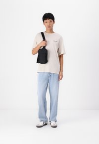 Svart nylon skulderveske med justerbar stropp, slankt design og glidelås, holdt av en person iført en lys beige T-skjorte og blå jeans.