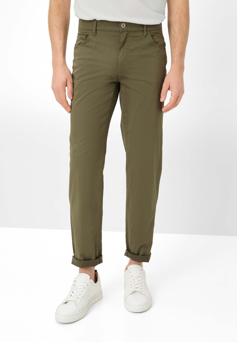 Mann trägt hochgekrempelte olivgrüne Hose und weiße Turnschuhe, steht vor einem schlichten weißen Hintergrund.