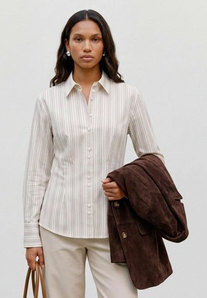 Femme aux longs cheveux foncés porte une chemise beige à rayures et un pantalon clair, tenant une veste marron dans un bras et la anse d’un sac marron dans l’autre.