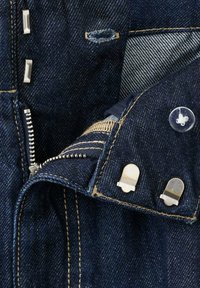 Primer plano de mezclilla azul oscuro con cremallera plateada, ojales, botón transparente y tachuelas metálicas en secciones de tela cosida.