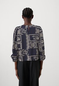 Blouse à motifs marins avec des designs floraux et géométriques blancs, col rond, manches bouffantes et une ouverture en forme de clé au dos.