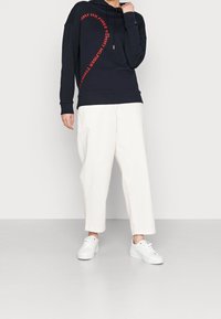 Granatowa bluza z kapturem z czerwonym logo "Tommy Hilfiger" w zaokrąglonym wzorze, zestawiona z białymi szerokimi spodniami i białymi sneakersami. Teksturowany materiał, luźny krój.