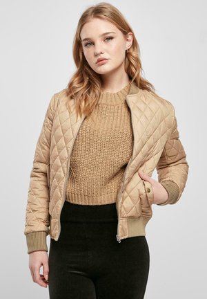 Nuori nainen, jolla on aaltoilevat vaaleat hiukset, hänellä on beige tikattu takki neulotun ruskean neuleen ja mustien housujen päällä, seisoo yksiväristä taustaa vasten.