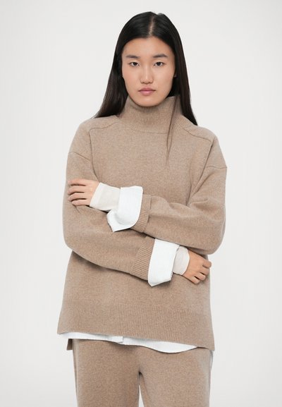 Vrouw met lang, donker haar in een beige gebreide coltrui, bijpassende broek en witte shirtmanchetten die haar armen over elkaar slaat tegen een effen achtergrond.