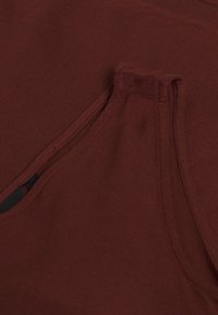 Bordowy materiał o gładkiej fakturze, z boczną kieszenią przeszytą pasującą nicią. Projekt zawiera delikatny połysk.