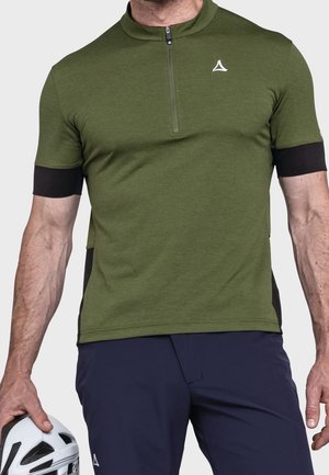 Mann trägt grünes kurzärmeliges Fahrradtrikot mit Reißverschluss und marineblaue Shorts, hält einen weißen Fahrradhelm in der linken Hand.