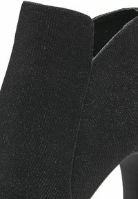 Czarna tkaninowa botka za kostkę o teksturowanej powierzchni, z delikatnymi akcentami brokatu i eleganckim, wysokim obcasem.