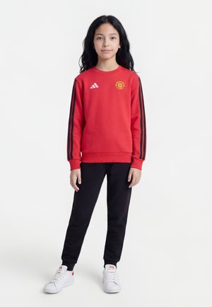 Copil în picioare, purtând un hanorac roșu Manchester United cu dungi negre și pantaloni negri, pantofi sport albi, pe un fundal alb simplu.