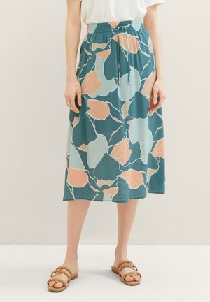 Femme portant une jupe midi fleurie bleu sarcelle, pêche et bleu clair avec une taille à cordon, un haut blanc et des sandales beige, debout contre un mur neutre.