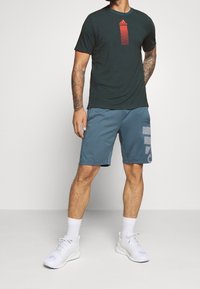 Tmavě zelené tričko s červenými vertikálními pruhy a logem Adidas, kombinované s modrými sportovními šortkami s grafickým potiskem a bílými teniskami.