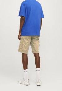 T-shirt in cotone blu, pantaloni cargo khaki con tasche laterali, sneaker bianche e calze bianche con logo nero. Sfondo neutro.