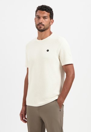 Kurzärmliges creamfarbiges Textshirt mit Rundhalsausschnitt, das ein kleines schwarzes sechseckiges Logo auf der Brust hat. Kombiniert mit beigen Hosen.