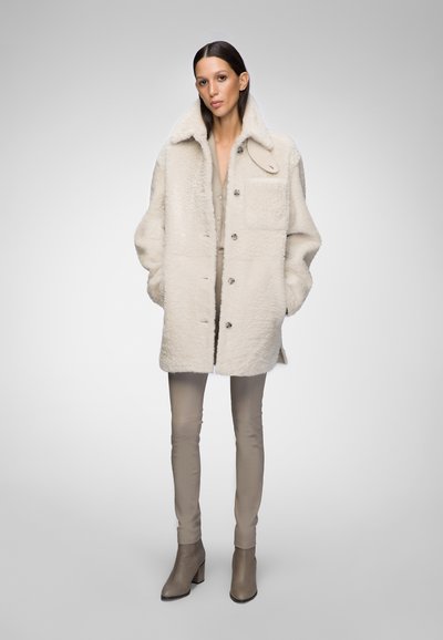 Cappotto in shearling bianco con ampio colletto e chiusura a bottone, dotato di tasca frontale e abbinato a pantaloni beige aderenti e stivaletti alla caviglia.