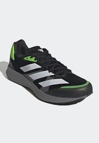 Scarpa sportiva nera con dettagli verde, superiore in rete, design a strisce bianche, suola testurizzata e marchio ben visibile sul tallone e sulla linguetta.