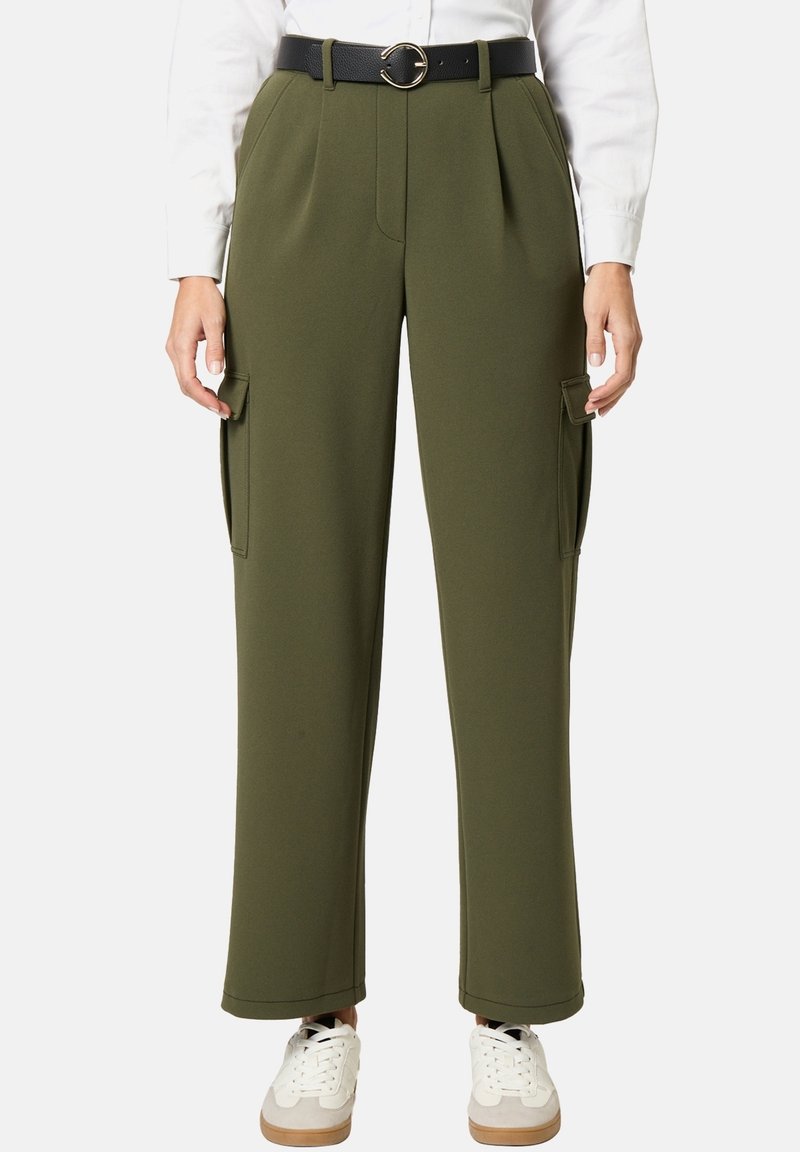 Femme portant un pantalon cargo ample vert olive avec poches à rabat, ceinture noire avec boucle ronde, chemise blanche et baskets blanches.