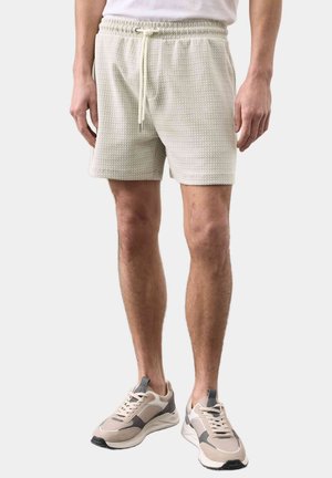 Man draagt crèmekleurige gebreide shorts met trekkoord en beige-grijze sneakers, staande tegen een effen lichte achtergrond.