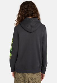 Persoon met krullend haar die een zwarte hoodie met groene bladranken op de mouwen en een bruine broek met donkere camouflageprint draagt, gezien van achteren.