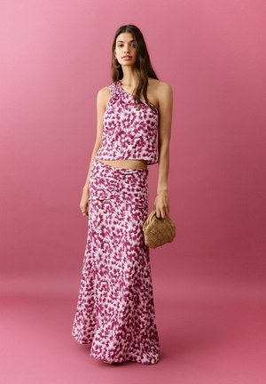 Ženska, ki nosi roza in belo cvetlično majico brez rokavov z enim ramenom ter ujemajočo dolgo krilo, drži pleteno torbico na roza ozadju.