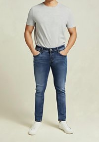 Man wearing a light gray short-sleeve t-shirt, blue slim-fit jeans, white slip-on sneakers, hands in pockets, standing on light background.