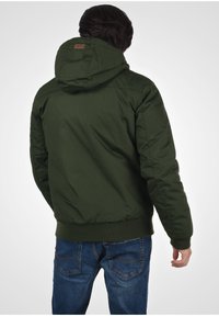 Giacca verde imbottita con cappuccio, polsini elastici e orlo. Presenta una patch in pelle con il logo sulla schiena. Indossata con jeans blu.