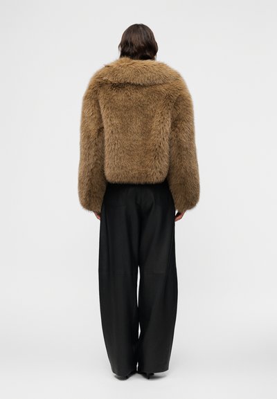 Άτομο που φοράει καφέ κοντό μπουφάν από συνθετική γούνα και χαλαρό μαύρο παντελόνι με φαρδιά μπατζάκια, στέκεται με την πλάτη προς τα έξω μπροστά σε απλό φόντο.