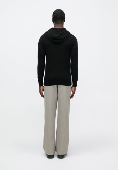 Homem de pé de costas, vestindo um hoodie preto, calças largas cinza claro e sapatos pretos, contra um fundo claro e simples.