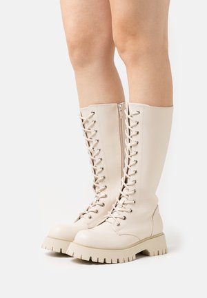 Jambes portant des bottes beiges à lacets montant jusqu'aux genoux avec semelles épaisses, photographiées sur un fond blanc uni.