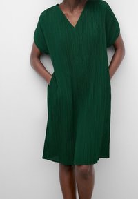 Kvinna i en knälång, kortärmad, plisserad mörkgrön klänning med V-ringning och händerna i sidorna som står framför en enfärgad bakgrund.