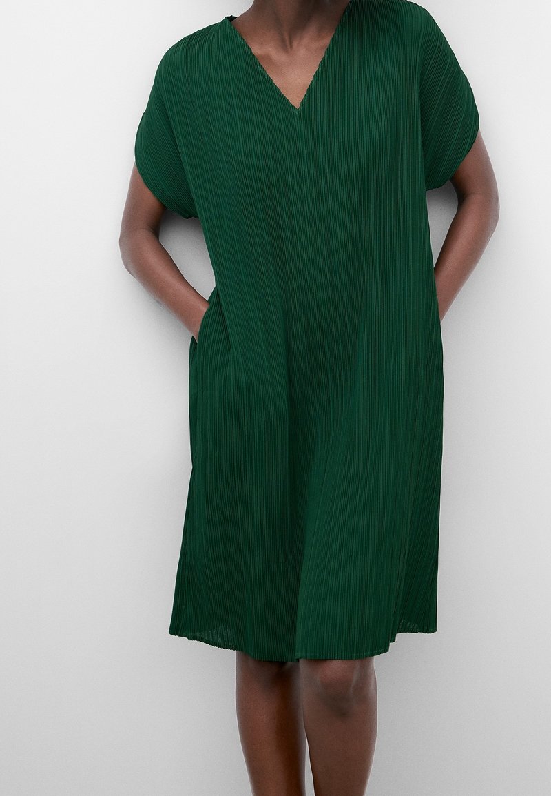 Kvinna i en knälång, kortärmad, plisserad mörkgrön klänning med V-ringning och händerna i sidorna som står framför en enfärgad bakgrund.