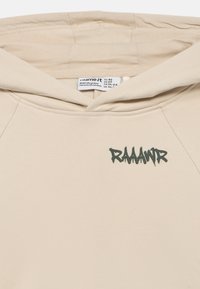 Μπεζ φούτερ με κουκούλα για νήπιο με την ένδειξη "RAAAWR" τυπωμένη με μαύρα γράμματα στο στήθος και μια εμφανή ετικέτα στο εσωτερικό του γιακά.
