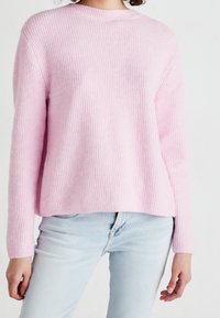 Osoba ubrana w jasnoróżowy sweter z prążkowanej wełny i jasnoniebieskie dżinsy na tle jednolitego białego tła.