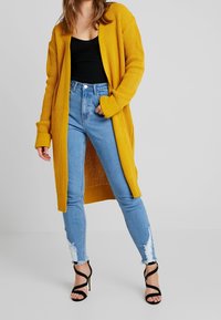 Cárdigan amarillo de punto con frente abierto, textura acanalada y mangas largas, llevado sobre una camiseta negra y combinado con jeans azul desgastados y tacones negros.