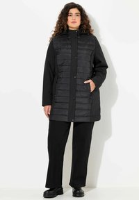Giacca nera isolante con parte anteriore trapuntata, bottoni a scatto, maniche lunghe e tasche; abbinata a pantaloni neri a gamba larga e stivali.