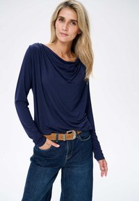 Navy-blaues Langarmoberteil mit drapiertem Ausschnitt, kombiniert mit dunkelblauen, hoch taillierten Jeans und einem beigen Gürtel mit einer dekorativen Schnalle.