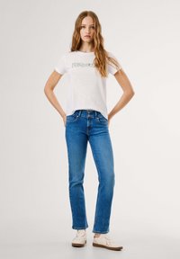 Ung kvinne med langt bølgete hår, iført en hvit t-skjorte med trykk, blå denimjeans og hvite joggesko, står med hendene på hoftene.