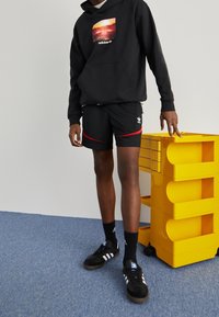 Felpa nera con stampa fotografica e logo, abbinata a pantaloni corti neri con righe rosse. Sneakers nere e calze completano l'outfit.