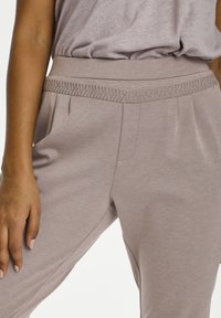 Nahaufnahme einer Person, die mauvefarbene Hose mit dekorativen Nähten am Bund und einem passenden mauvefarbenen Oberteil trägt, das in die Hose gesteckt ist.