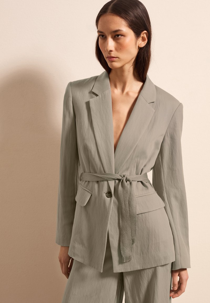 Lichtgroene blazer met een diepe V-hals, een ceintuur in de taille en twee voorzakken. De stof lijkt gestructureerd en is geschikt voor casual of formele kleding.