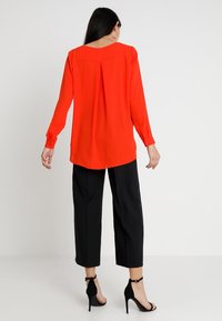 Blouse rouge à manches longues avec un pli dans le dos, portée avec un pantalon noir raccourci et des sandales à talons hauts noires sur un fond neutre.