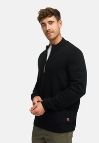 Mustapohjainen vetskariaukko cardigan, jossa on korkea kaulus, valmistettu teksturoidusta kankaasta. Ominaisuuksiin kuuluu huomaamaton bränditag alhaalla vasemmalla ja resorit hihoissa.